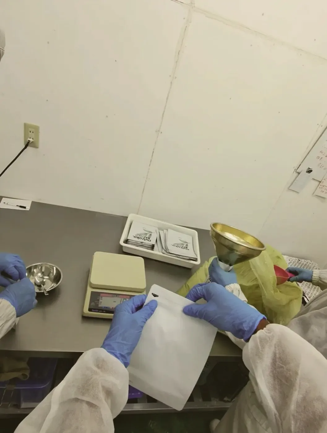 Holding a pouch open at the filling station, with a brass funnel, bench scale, and a tray of Nutroko labels on the table