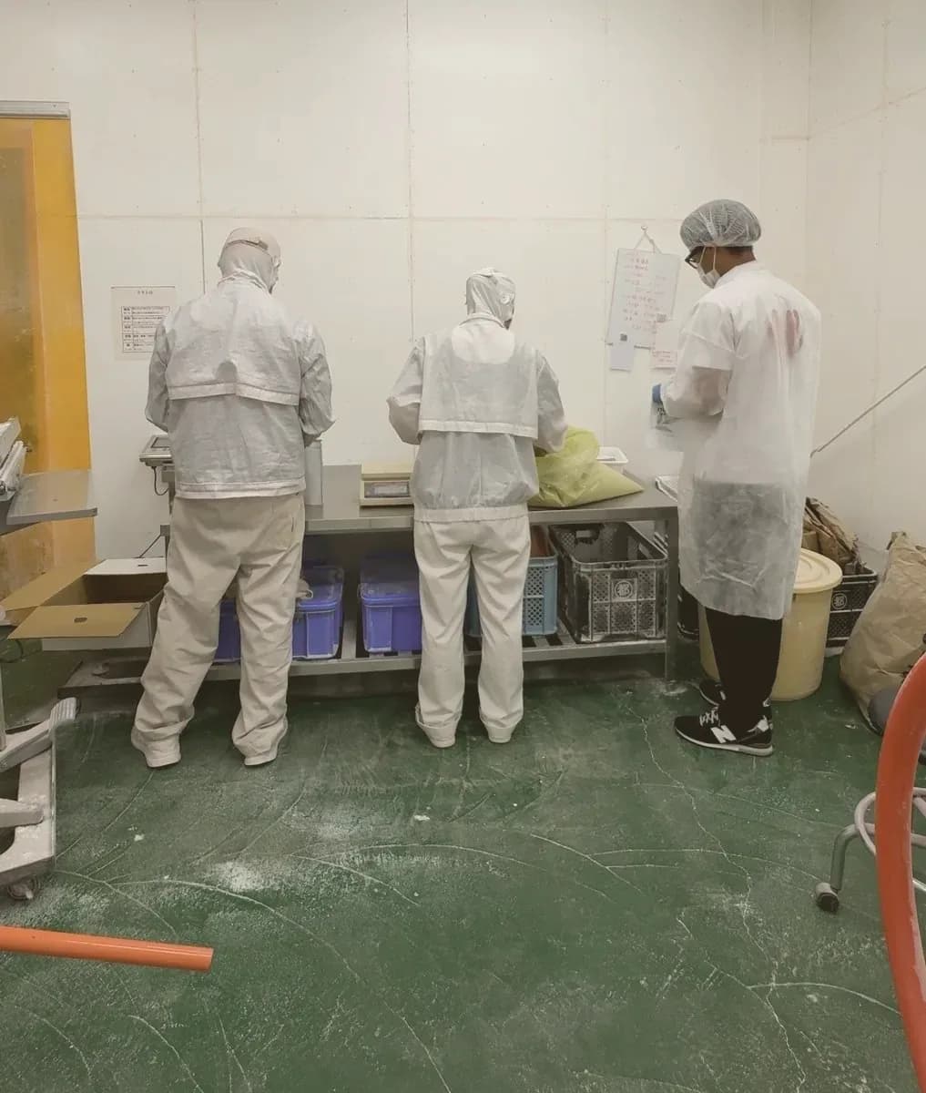 Minami and two others in food-grade whites weighing and sealing Nutroko pouches at a steel table in the Osaki-cho sweet potato factory