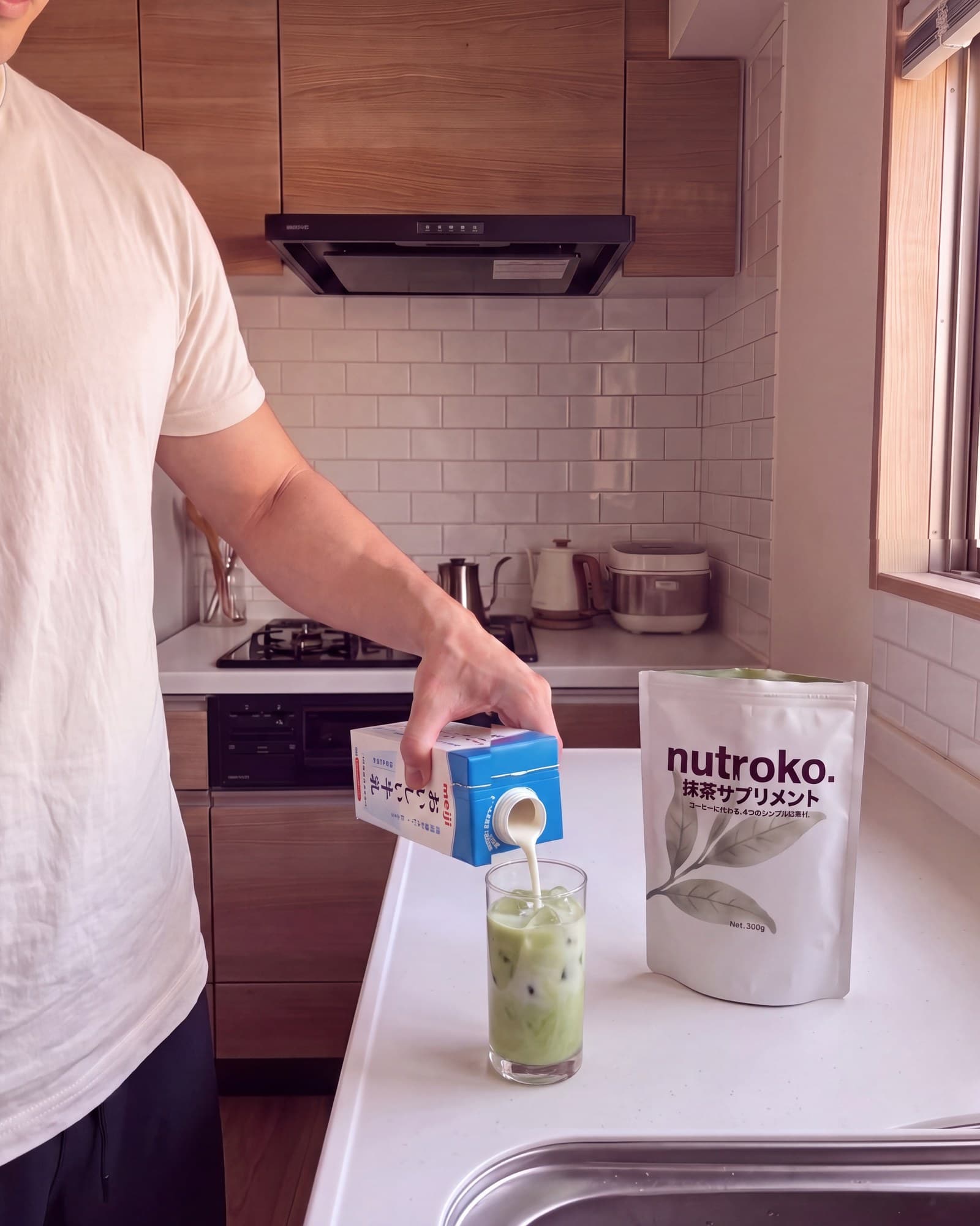 Pouring milk from a Japanese carton into a tall glass of prepared Nutroko matcha supplement over ice, the pouch standing beside it on the kitchen counter