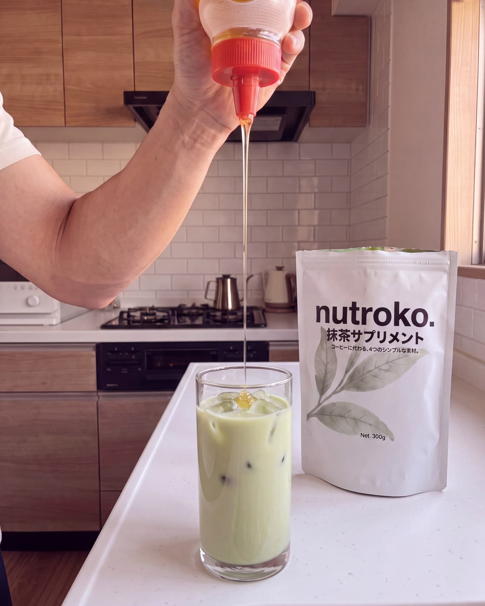 Drizzling honey into a tall glass of prepared Nutroko matcha supplement over ice, the pouch standing beside it on the kitchen counter
