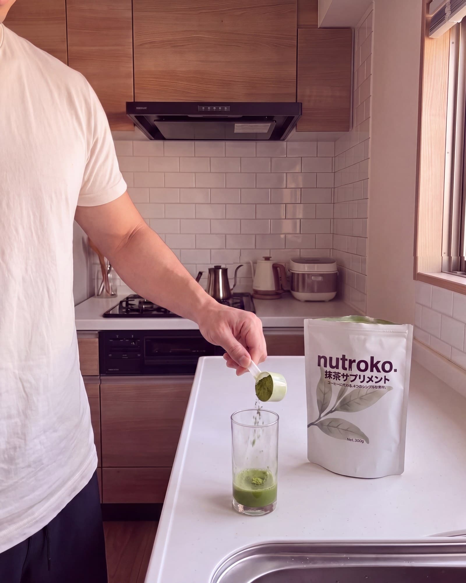 Tipping a scoop of green Nutroko powder into a tall glass on a kitchen counter, the pouch standing beside it